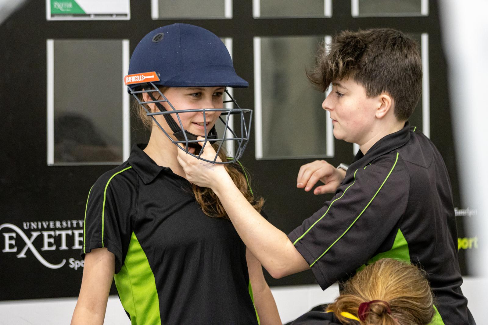 Newton Abbot Young Leader, Libby, helps a participant with putting on the kit for her first go at hardball cricket.